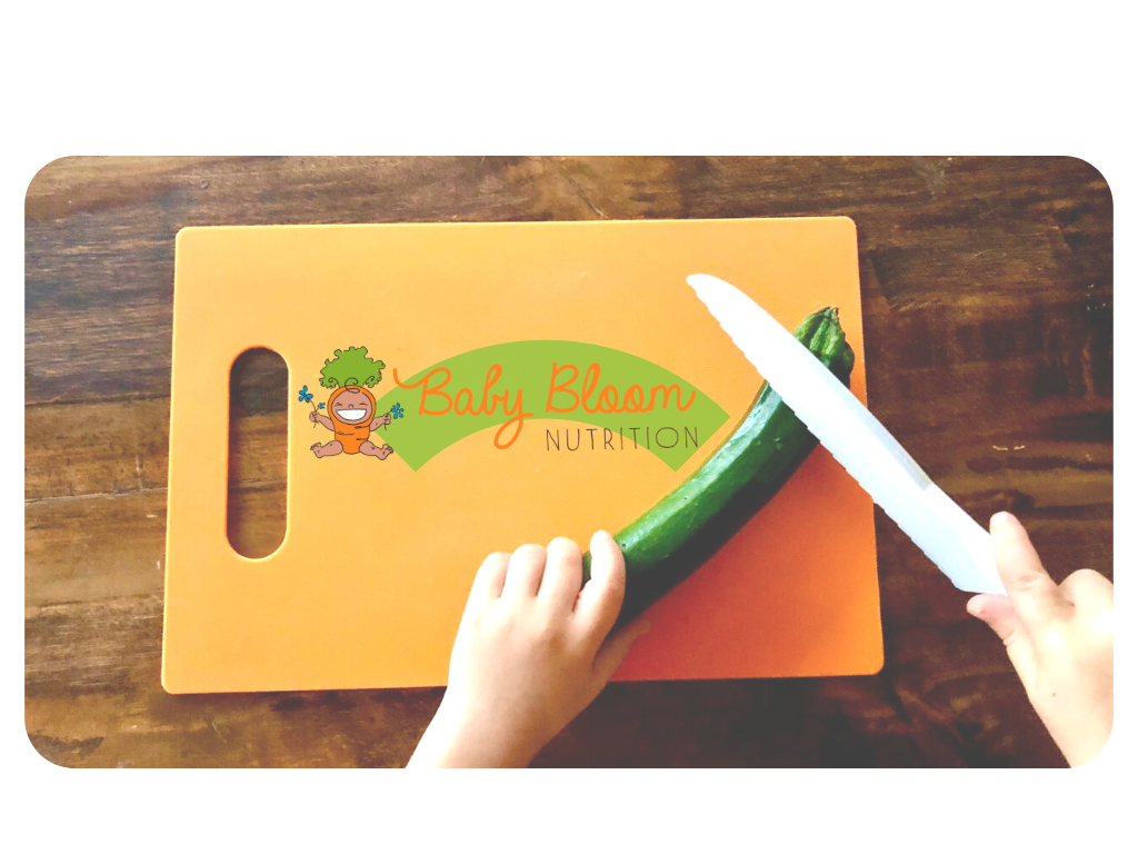 orange cutting board on dark wooden table, Baby Bloom Nutrition carrot baby logo, a child with a white nylon knife cutting a zucchini